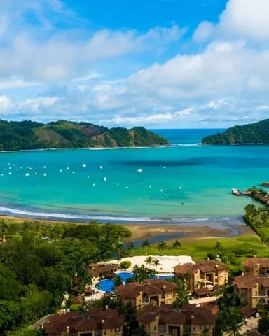 Aerial view of turquoise bay with crescent beach, forested headlands, marina, and resort buildings below