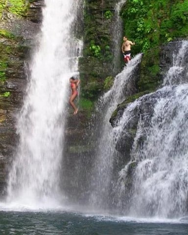 Two people swimming in a pool beneath multiple cascading waterfalls surrounded by moss-covered rock cliffs and green vegetation