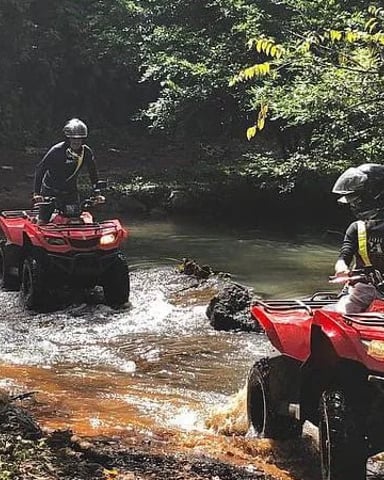 Two people on red ATVs crossing a shallow stream through dense jungle vegetation