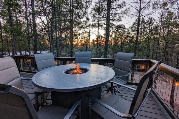Zwei moderne Hütten im Wald: Links eine erhöhte Terrasse mit Treppe und Geländer in der Abenddämmerung; rechts ein Sitzplatz mit Essmöbeln und Pendelleuchten.